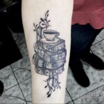 Фото тату на руке с изображением стопки книг, чашки кофе и веток с листьями. Татуировка выполнена в черно-белой гамме.