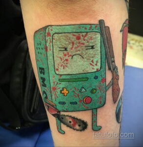 Фотография татуировки с изображением персонажа BMO из мультфильма Adventure Time, покрытого брызгами крови, выполненной на руке в ярких цветах.