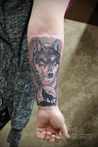 Фото тату волка, воющего на луну, на предплечье. Черно-серый рисунок татуировки в стиле реализм с детальной проработкой шерсти и лунного пейзажа.