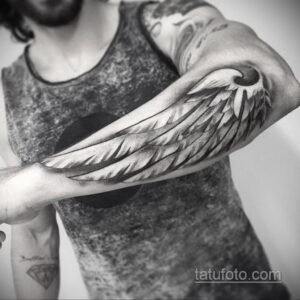 Фото татуировки крыла на руке мужчины. Детализированное изображение перьев в черно-серых тонах, занимает большую часть предплечья.