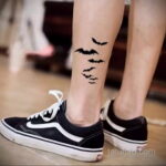 Фото тату: маленькие черные летучие мыши, летящие вдоль лодыжки. Изящный рисунок татуировки для девушек, выполненный в минималистичном стиле.