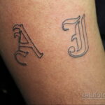Фото тату с инициалами 'A' и 'J', выполненное в черно-серых тонах на запястье. Изящный шрифт с тонкими линиями.