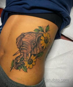 Фото татуировки головы слона, окруженной подсолнухами на боку. Рисунок татуировки выполнен в реалистичном стиле с использованием ярких цветов.