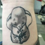 Фото тату: Реалистичное изображение спящего слоненка на руке, выполненное в черно-серых тонах. Детализированная татуировка с использованием теней и полутонов.