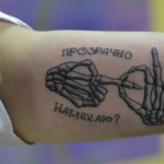 Черно-белое фото татуировки на руке с изображением скелетических рук, держащихся за пальцы. Надпись 'Прозрачно намекаю?' сверху и снизу.