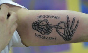 Черно-белое фото татуировки на руке с изображением скелетических рук, держащихся за пальцы. Надпись 'Прозрачно намекаю?' сверху и снизу.