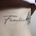 Фото тату на запястье: надпись 'Familia' выполненная красивым шрифтом, рядом изображен маленький цветок, черно-серые тона.