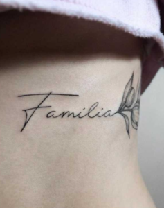 Фото тату на запястье: надпись 'Familia' выполненная красивым шрифтом, рядом изображен маленький цветок, черно-серые тона.