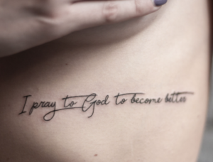 Фото татуировки надпись на боку. Черная надпись 'I pray to God to become better' выполнена каллиграфическим шрифтом. Минималистичный рисунок татуировки.