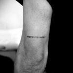 Фото минималистичного тату с надписью "Memento Mori" на руке. Тонкая черная надпись, выполненная простым шрифтом.