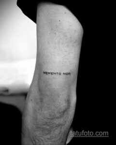 Фото минималистичного тату с надписью "Memento Mori" на руке. Тонкая черная надпись, выполненная простым шрифтом.