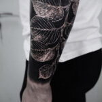 Фото тату с изображением ветки с листьями, выполненной в технике blackwork на руке мужчины. Детализированный рисунок с использованием только черного цвета.