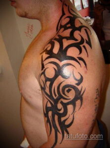 Фото тату: классическая татуировка в стиле трайбл, выполненная в черном цвете на плече и верхней части руки мужчины.
