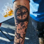Фото тату на предплечье: компас, карта и надпись "Not all who wander are lost". Детализированный рисунок в стиле blackwork.