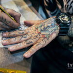 Фото мастера тату, рисующего эскиз татуировки на руке клиента, с множеством эскизов на заднем плане.