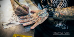 Фото мастера тату, рисующего эскиз татуировки на руке клиента, с множеством эскизов на заднем плане.