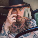 Фотография мужчины с множеством татуировок в стиле олд скул на руках, груди и шее. Изображены змеи, цветы, надписи и другие традиционные элементы.