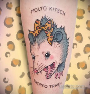 Фото татуировки с изображением опоссума в бантике и надписью "Molto Kitsch Troppo Trash", выполненной в ярких цветах на руке.