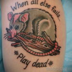 Фото тату опоссума, притворяющегося мертвым, с надписью 'When all else fails... Play dead' на руке. Яркий и красочный рисунок в стиле традиционной татуировки.
