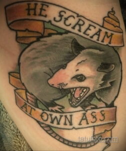 Фото тату злого опоссума с надписью 'He scream at own ass' в стиле олд скул на плече. Черно-серые тона и детализированная проработка.