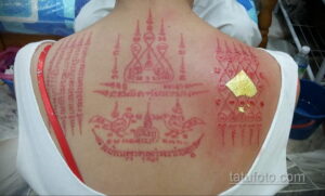 Фото тату: Татуировка на спине с тайскими символами, выполненными красной краской, и золотой наклейкой. Вид сзади.