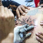 Фото тату: мастер наносит сакральную татуировку на спину клиента. Видны руки мастера, тату-машинка и уже нанесенные символы.