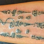 Фото тату: черно-серая татуировка с тайскими символами и цифрами на предплечье. Видны следы заживления и небольшие повреждения.