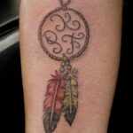 Фото тату: рисунок ловца снов с буквами внутри круга на женском предплечье. Эскиз татуировки выполнен в черно-серых тонах.