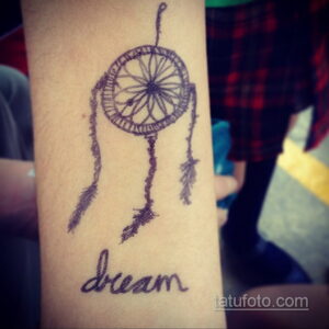 Фото минималистичной татуировки ловца снов на руке с надписью "dream". Изображение выполнено в черно-серых тонах.