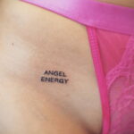 Фото тату надписи 'Angel Energy' выполненной черными чернилами на коже. Минималистичный рисунок татуировки расположен на груди под розовым бельем.