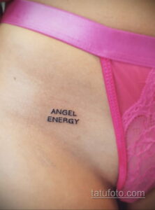 Фото тату надписи 'Angel Energy' выполненной черными чернилами на коже. Минималистичный рисунок татуировки расположен на груди под розовым бельем.