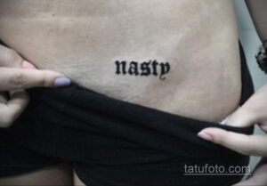 Фото тату надписи 'nasty' черным цветом, выполненной простым шрифтом на коже талии девушки. Минималистичный рисунок татуировки.
