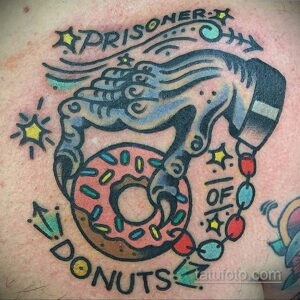 Фото тату: рука, держащая пончик, в стиле олд скул с надписью "Prisoner of Donuts". Винтажный рисунок татуировки.