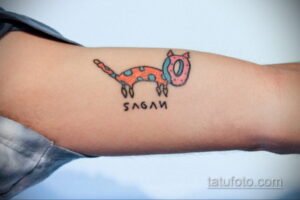 Фото тату стилизованного кота с надписью "SAGAN" на руке. Минималистичный и оригинальный рисунок татуировки в ярких цветах.