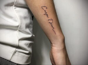 Фотография татуировки с надписью 'Carpe Diem', выполненной элегантным шрифтом на внутренней стороне предплечья. Черно-белый дизайн.
