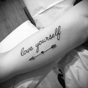 Фото татуировки с надписью 'love yourself', стилизованной под рукописный шрифт, и стрелой, пронзающей сердце. Черно-белый дизайн на предплечье.