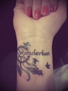 Фото тату: минималистичный ловец снов с летящими птицами и надписью 'Wanderlust' на запястье.
