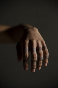 Фото минималистичной татуировки на руке, состоящей из тонких черных линий, расположенных вокруг пальцев. Современный рисунок татуировки в стиле line work.