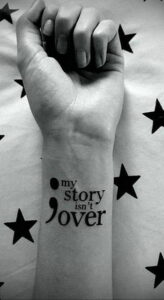 Фото тату: вдохновляющая надпись 'my story isn't over' с точкой и запятой на запястье девушки.
