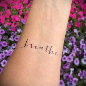 Фото тату: изящная надпись 'breathe' в виде пульсирующей линии на запястье девушки на фоне цветущих растений.