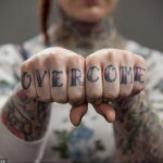 Фото тату на костяшках пальцев с надписью 'OVERCOME' черными чернилами. Руки с татуировками на предплечьях.