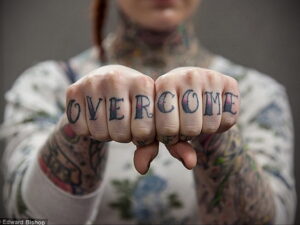 Фото тату на костяшках пальцев с надписью 'OVERCOME' черными чернилами. Руки с татуировками на предплечьях.