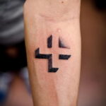 Фото минималистичной татуировки в виде геометрического креста, расположенного на запястье. Рисунок татуировки выполнен в черном цвете с четкими линиями.