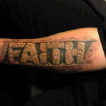 Фото тату на руке с надписью "FAITH", выполненной в виде высеченных в камне букв. Реалистичный рисунок татуировки.