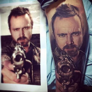 Фото тату на руке с реалистичным портретом Джесси Пинкмана из сериала Breaking Bad с пистолетом в руке. Детализированный рисунок татуировки.