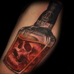 Фото тату на предплечье: реалистичная бутылка с жидкостью, внутри которой виден череп. Темные тона, детализированная проработка.