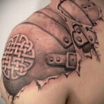 Фото тату в стиле blackwork, изображающего плечо в рыцарских доспехах с кельтским узором на наплечнике. Рисунок выполнен в черных тонах.