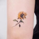 Фото тату на запястье: реалистичное изображение цветка подсолнуха с желтыми лепестками и зелеными листьями, выполненное в акварельном стиле.