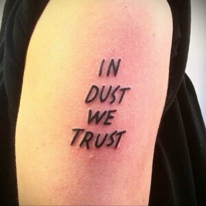 Фото тату на руке: фраза "In Dust We Trust" написана черным шрифтом в стиле леттеринг, с неровными краями.
