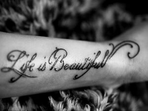 Фото татуировки на запястье с надписью "Life is Beautiful", выполненной каллиграфическим шрифтом. Тату выполнено в черном цвете.